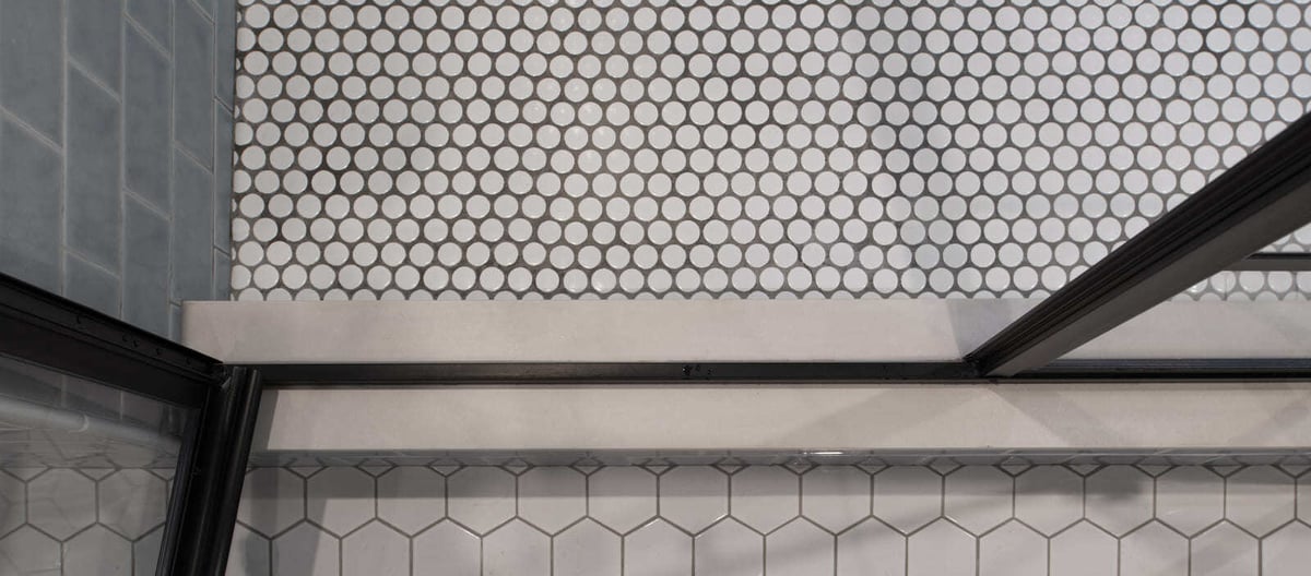 Close-up of a luxurious shower threshold with black metal framing, white hexagon floor tiles, and sleek gray subway walls in an Acton, MA remodel